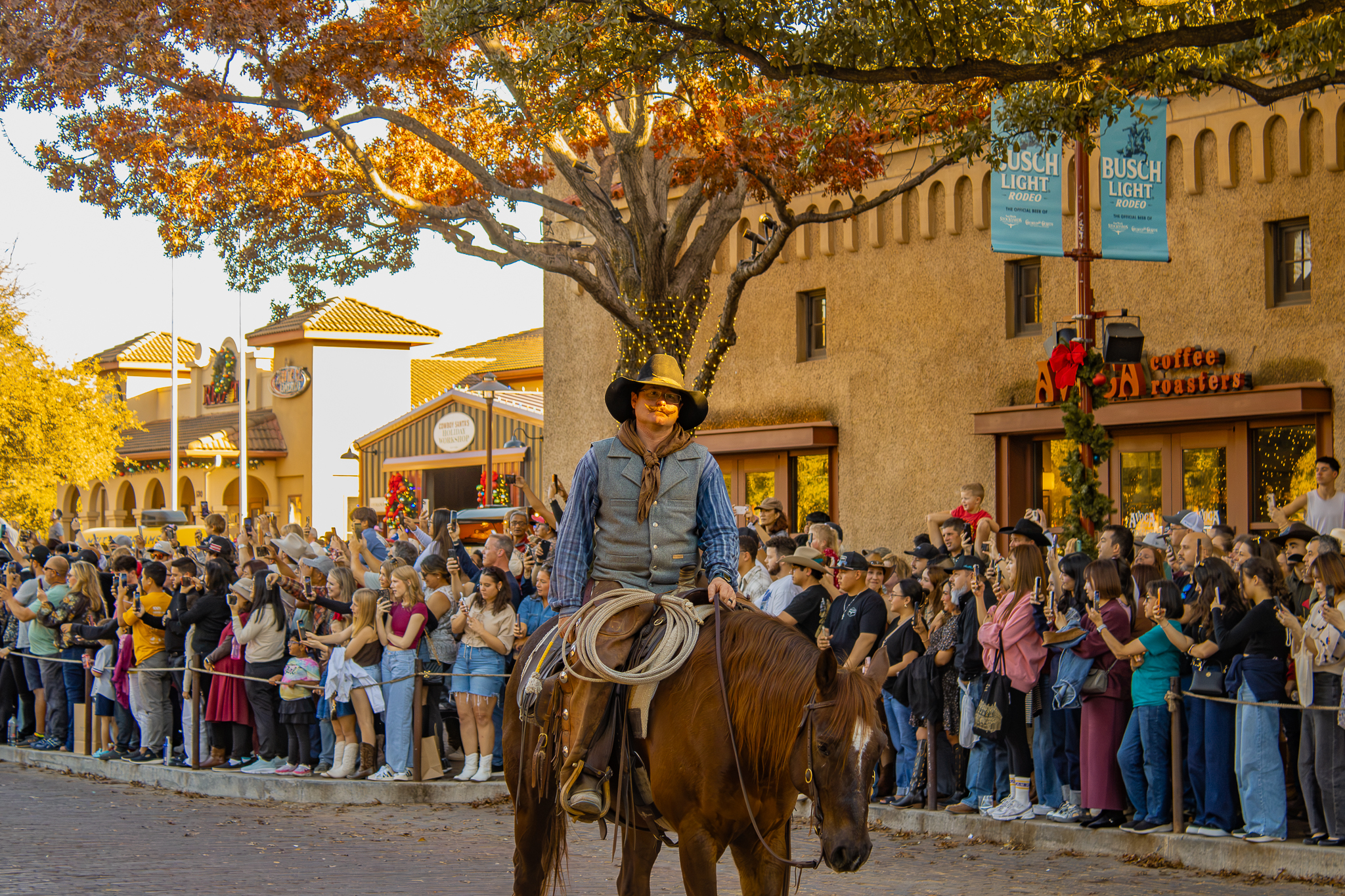 A rider in a wide-brimmed hat at the golden hour, a crowd and stone storefronts behind him.